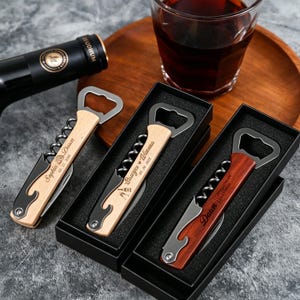 Op de afbeelding: Drie multifunctionele wijnopeners met houten handvatten in zwarte geschenkdozen. Elk hulpmiddel bevat een metalen kurkentrekker, een flesopener en een foliesnijder. De handvatten zijn gegraveerd met namen en data: Sophia & Dawn, 03.11.2026, Sawyer & Winona, 05.20.2025 en Dawn, 21.11.2025.