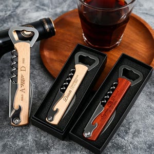 May include: Three waiter-style corkscrews with bottle openers, featuring wooden handles engraved with names and dates. Two are in black gift boxes, and one is loose. The handles are light and dark wood, with silver-toned metal mechanisms.
