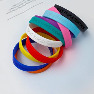 Peut inclure: Une collection de bracelets en silicone colorés empilés les uns sur les autres. Les bracelets sont noirs, roses, bleu clair, violets, sarcelle, blancs, jaunes, rouges, bleus et orange. Chaque bracelet porte le texte Best Friends Together en relief.