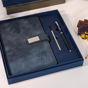Puede incluir: Un juego de regalo azul marino en una caja, que incluye un diario de cuero con un cierre plateado, un bolígrafo y un lápiz óptico. El diario está grabado con las iniciales "V E" y el nombre "Victor Orona". El juego se presenta en una caja azul oscuro.