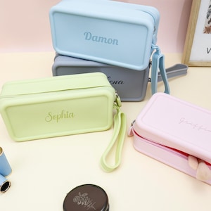 以下が含まれることがあります： パステルカラーの長方形の化粧ポーチのコレクション。色はライトブルー、グレー、グリーン、ピンク。各ポーチにはリストストラップと名前（Damon、Lena、Sophia、Griffin）がプリントされています。ポーチは柔らかくマットな素材で作られています。