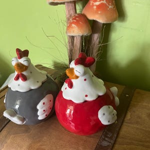 Peut inclure: Deux récipients en céramique en forme de poule, un gris et un rouge, avec des accents blancs et des pois rouges. Les poules ont des crêtes rouges et des becs jaunes. Derrière, des ornements décoratifs en forme de champignons orange et blancs.