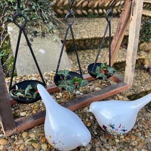 Può includere: Una cornice in legno rustico con tre fioriere sospese in metallo nero, ognuna con vegetazione e bacche rosse. Due figurine di uccelli in ceramica bianca con disegni floreali sono in primo piano.