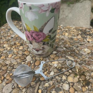 Peut inclure: Une tasse en céramique avec un visage de lapin et un motif floral, accompagnée d'une boule à thé en acier inoxydable. La tasse présente un fond vert clair avec des fleurs roses et blanches. L'infuseur à thé est argenté et possède une boule en maille.