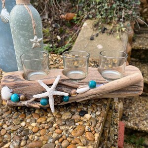 Peut inclure: Un bougeoir en bois flotté avec trois photophores en verre transparent. Le support est décoré d'une étoile de mer blanche, de coquillages et de perles turquoise. Deux bouteilles décoratives sont en arrière-plan.