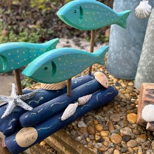 Peut inclure: Trois sculptures de poissons en bois turquoise avec des détails de points blancs, montées sur une base en bois flotté bleu. La base est décorée de coquillages et d'une étoile de mer blanche. Thème côtier décoratif.