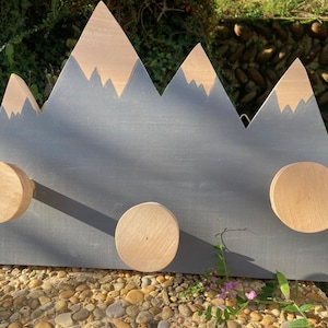 Può includere: Appendiabiti a parete a forma di montagna in legno, con base grigia e dettagli in legno naturale. Le cime delle montagne sono marroni chiare e sono presenti tre pomelli rotondi in legno per appendere oggetti. Adatto per una cameretta.