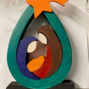 May include: A wooden nativity scene with a teal teardrop frame, featuring Mary, Joseph, and baby Jesus. The figures are painted in blue, brown, red, and orange, with a bright orange star at the top. The base is black.