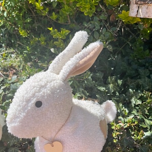 May include: A plush white bunny rabbit with a heart-shaped wooden tag around its neck. The bunny has tan inner ears and accents on its rear. The soft toy is set against a backdrop of green foliage and a small birdhouse.