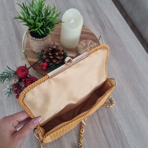 Op de afbeelding: Een mosterdgele gebreide clutch met een gouden kettingband en een gouden sluiting. De tas is open en de binnenkant is gevoerd met een lichtbruine stof.