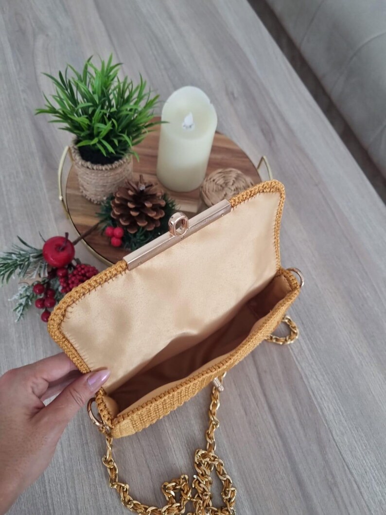 Op de afbeelding: Een mosterdgele gebreide clutch met een gouden kettingband en een gouden sluiting. De tas is open en de binnenkant is gevoerd met een lichtbruine stof.