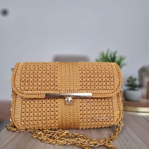 Op de afbeelding: Een mosterdgele gebreide handtas met een gouden kettingband en een gouden sluiting. De tas heeft een "Handmade" label aan de voorkant.