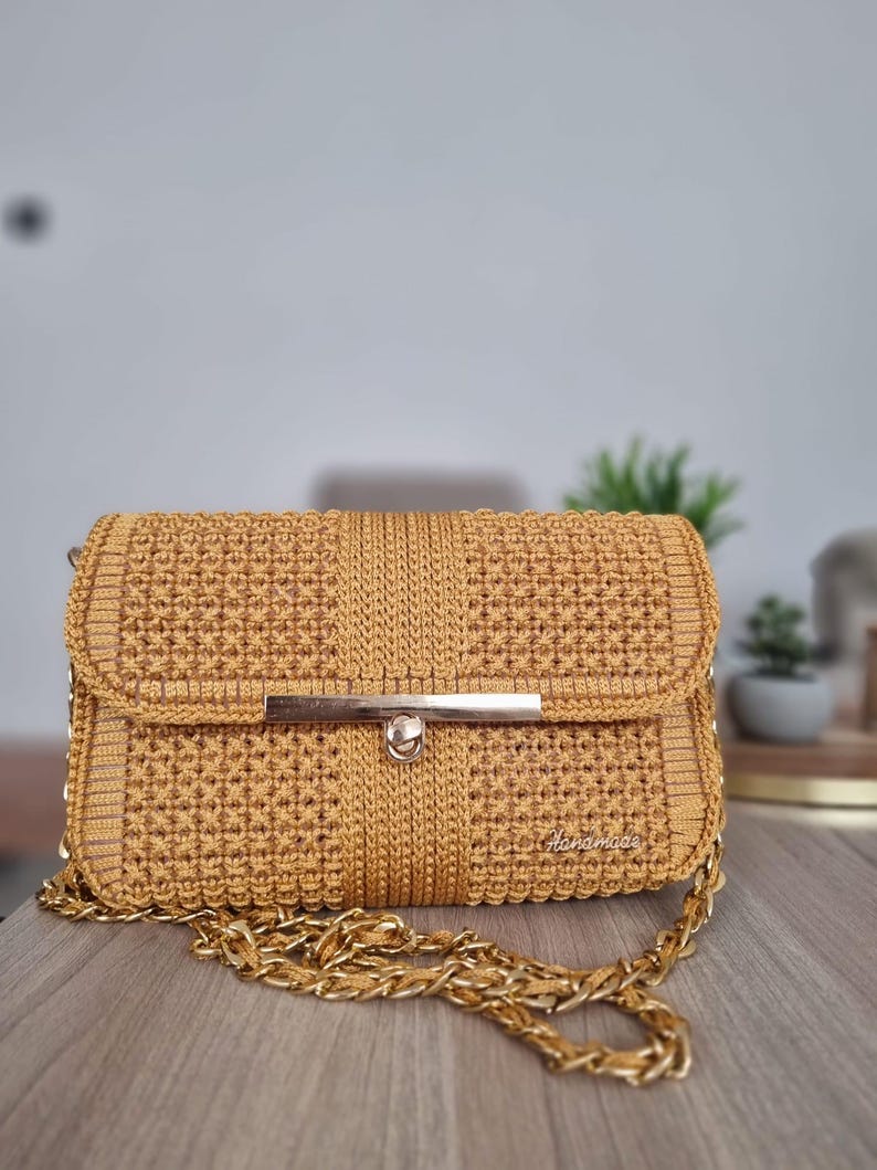 Op de afbeelding: Een mosterdgele gebreide handtas met een gouden kettingband en een gouden sluiting. De tas heeft een "Handmade" label aan de voorkant.