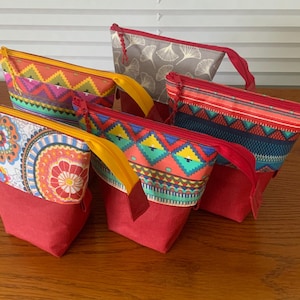 Könnte beinhalten: Fünf farbenfrohe Reißverschlusstaschen mit roten Böden. Die Taschen weisen lebendige Muster auf, darunter geometrische und florale Designs, mit gelben und roten Reißverschlussanhängern. Die Taschen sind auf einer Holzoberfläche angeordnet.