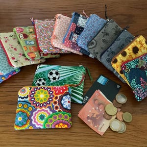Könnte beinhalten: Eine Sammlung von 12 bunten Reißverschlusstaschen mit verschiedenen Mustern, darunter Eulen, Blumen und geometrische Designs. Die Taschen sind in einem Halbkreis auf einer Holzoberfläche mit Münzen und einer Kreditkarte angeordnet.