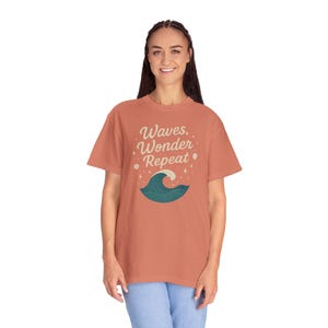 Op de afbeelding: Koraalrood T-shirt met de tekst "Waves, Wonder, Repeat" in een speels lettertype. Onder de tekst staat een gestileerde golf in turquoise en wit. Het shirt is een relaxte, casual stijl.