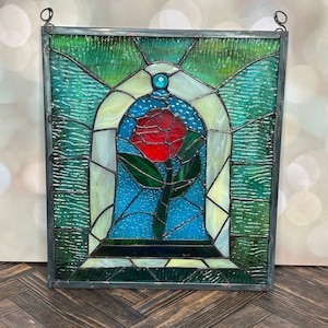 May include: Stained glass panel featuring a red rose within a blue and white arched enclosure. The panel is framed with green and clear glass, and has a metal border with hanging loops. The rose has green leaves and a long stem.
