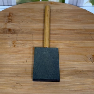 Op de afbeelding: Een donkergrijze slijpsteen met een rechthoekige vorm en een houten handvat. De handgreep is lichtbruin en bevestigd aan de bovenkant van de steen. De steen ligt op een houten oppervlak.