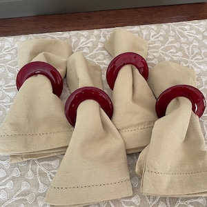 Pode incluir: Quatro guardanapos de linho bege com argolas de guardanapo de vidro vermelho. As argolas têm um design simples e arredondado.