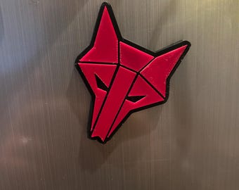 Może przedstawiać: Geometryczny magnes w kształcie głowy wilka o żywym czerwonym kolorze, z ostrymi, kanciastymi liniami i wyraźną czarną obwódką. Stylizowany pysk zwierzęcia składa się z wyraźnych trójkątnych i trapezoidalnych segmentów.
