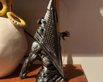 Puede incluir: Una escultura detallada y gris oscuro de una figura de Pyramid Head de la serie de videojuegos Silent Hill. La figura posa con su icónico casco en forma de pirámide y una gran cuchilla. La escultura se exhibe en una estantería de madera.