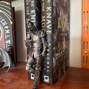 May include: A metallic silver-toned knight figurine standing between two books. The knight is wearing full plate armour and holding a small open book in its hands. The books are titled The Knight and the Moon and The Knave and the Moon.