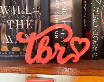 Puede incluir: Un letrero decorativo de color coral brillante que presenta las letras "TBR" en una fuente cursiva y fluida. El diseño incluye una forma de corazón integrada en la última letra, creando una línea continua y conectada en toda la palabra.