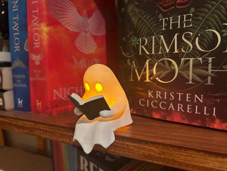 Puede incluir: Una luz de libro con forma de fantasma naranja brillante leyendo un peque&ntilde;o libro negro, colocada en una estanter&iacute;a de madera. El fantasma tiene dos ojos amarillos y una parte inferior blanca y fluida. Libros con t&iacute;tulos como "The Crimson Moth" est&aacute;n en el fondo.