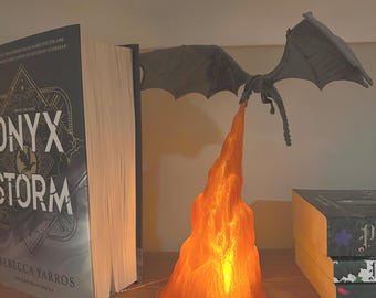 Puede incluir: Una figura de dragón gris con las alas extendidas, que parece exhalar un chorro de luz naranja. El dragón está colocado encima de libros en una estantería de madera. La luz ilumina al dragón y el área circundante.