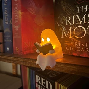 Puede incluir: Una luz de libro con forma de fantasma amarillo brillante leyendo un libro peque&ntilde;o, con una parte inferior blanca. La luz est&aacute; sobre una estanter&iacute;a de madera con libros. La luz del libro tiene dos luces en forma de ojo y un libro peque&ntilde;o.