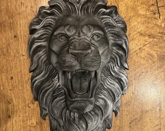 Puede incluir: Una escultura de pared con forma de cabeza de león, con un acabado metálico gris oscuro y texturas en capas visibles. El león se muestra con una melena abundante y fluida, con la boca abierta que deja ver los dientes y la lengua en relieve.