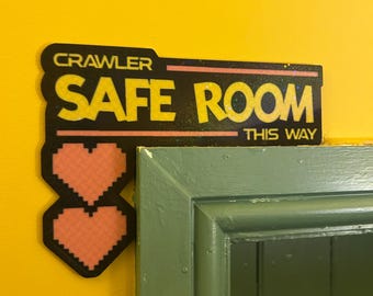 Może przedstawiać: Czarny prostokątny znak z żółtym napisem CRAWLER SAFE ROOM THIS WAY, z dwoma ułożonymi w stos pikselowymi różowymi sercami po lewej stronie. Znak jest zamontowany na żółtej ścianie nad rogiem zielonej ościeżnicy drzwiowej.