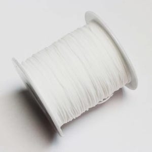 Può includere: Una bobina bianca di cordino sottile e flessibile avvolto strettamente attorno a un cilindro di plastica. Il cordino ha una consistenza liscia e uniforme, che copre l'intera lunghezza della bobina da un'estremità circolare all'altra, creando un aspetto bianco solido.
