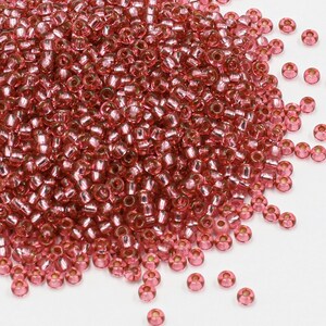 Peut inclure: Un grand tas de petites perles de verre rondes et roses avec un éclat métallique. Les perles sont éparpillées sur une surface blanche.