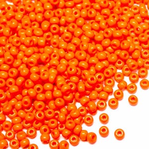 Könnte beinhalten: Nahaufnahme eines Haufens kleiner, runder, orangefarbener Perlen. Jede Perle hat ein Loch in der Mitte. Die Perlen sind auf einer weißen Oberfläche verstreut und erzeugen einen strukturierten Effekt. Die Perlen sind leuchtend orange.