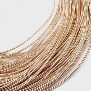 May include: A coiled length of metallic rose gold wire featuring a fine, textured, and ribbed surface. The wire is arranged in a series of concentric loops, creating a dense, uniform pattern of thin, flexible strands that catch the light.