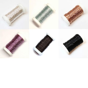 Op de afbeelding: Zes spoelen metallic garen in verschillende kleuren, waaronder zilver, groen, koper, paars, bruin en zwart. Elke spoel is gewikkeld op een witte plastic spoel, geschikt voor naai-, borduur- of knutselprojecten.