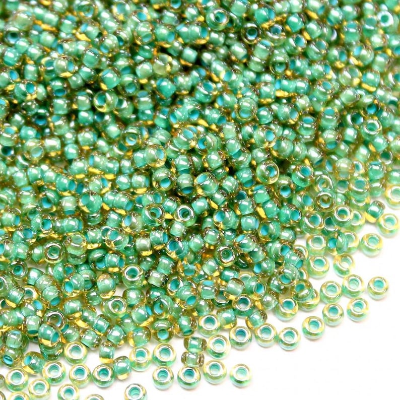 May include: A large pile of small, round, green glass beads with a gold iridescent finish. The beads are scattered on a white surface.