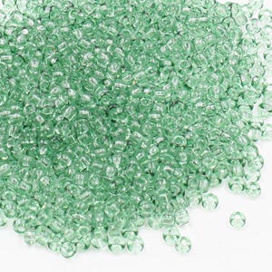 May include: A large pile of small, light green glass seed beads. The beads are round and have a slightly iridescent finish.