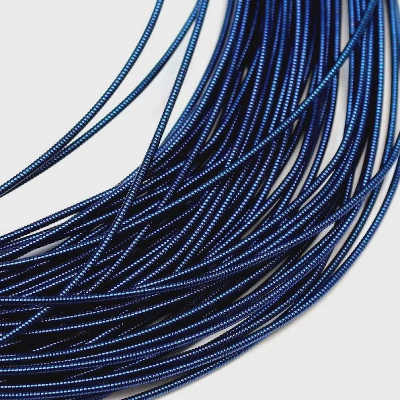 May include: A coil of flexible, deep blue metallic wire featuring a continuous, tightly wound spiral texture. The wire is arranged in multiple overlapping loops, creating a dense, uniform pattern of dark blue ridges and reflective surfaces.