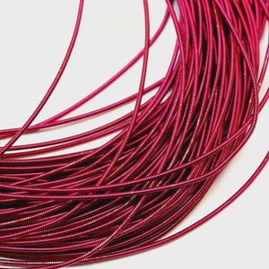 May include: A coil of flexible, metallic fuchsia wire featuring a continuous, tightly wound spiral texture. The thin, vibrant pink cord is arranged in overlapping loops, showcasing a consistent, ribbed surface throughout the entire length.