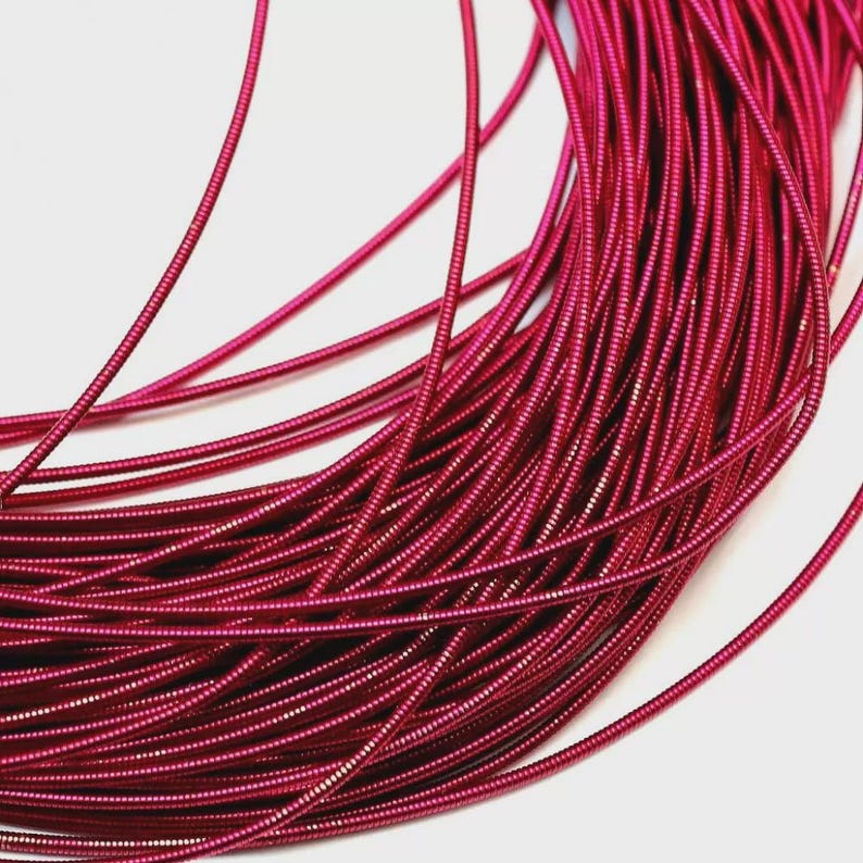 May include: A coil of flexible, metallic fuchsia wire featuring a continuous, tightly wound spiral texture. The thin, vibrant pink cord is arranged in overlapping loops, showcasing a consistent, ribbed surface throughout the entire length.