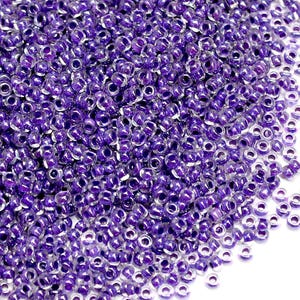 Puede incluir: Primer plano de un montón de pequeñas cuentas redondas moradas. Las cuentas tienen una calidad translúcida, con una superficie brillante y reflectante. Las cuentas están densamente empaquetadas, creando una exhibición texturizada y llamativa. Ideal para la fabricación de joyas y manualidades.