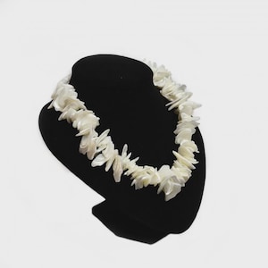 May include: A white shell necklace displayed on a black bust. The necklace is composed of irregularly shaped, off-white shell pieces, creating a textured and organic appearance. The necklace is presented against a plain white background.