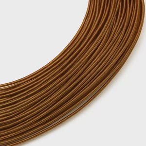 May include: A coil of metallic bronze elastic cord arranged in a curved, layered pattern. The cord features a tightly wound, ribbed texture that creates a consistent, repeating linear appearance across the entire length of the material.
