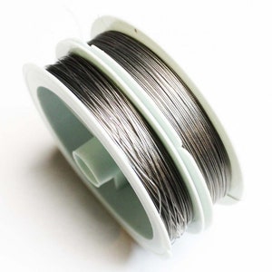 May include: A spool of thin, dark metallic wire wound tightly around a white plastic bobbin. The wire has a smooth, reflective surface and is coiled in multiple layers across the width of the spool, which features a central cylindrical core.