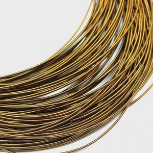 May include: A coiled length of metallic gold-colored wire featuring a distinct, tightly wound spiral texture along its entire surface. The wire is arranged in multiple overlapping circular loops, creating a dense, uniform pattern of reflective, textured golden strands.