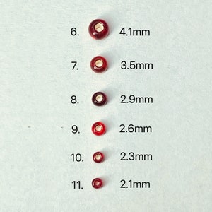 May include: Six red glass beads of varying sizes, ranging from 4.1 mm to 2.1 mm in diameter. The beads are arranged in a vertical line, with the largest bead at the top and the smallest bead at the bottom. The beads are all round and have a smooth, glossy finish.