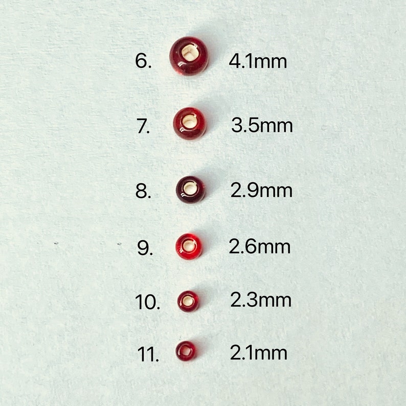 May include: Six red glass beads of varying sizes, ranging from 4.1 mm to 2.1 mm in diameter. The beads are arranged in a vertical line, with the largest bead at the top and the smallest bead at the bottom. The beads are all round and have a smooth, glossy finish.