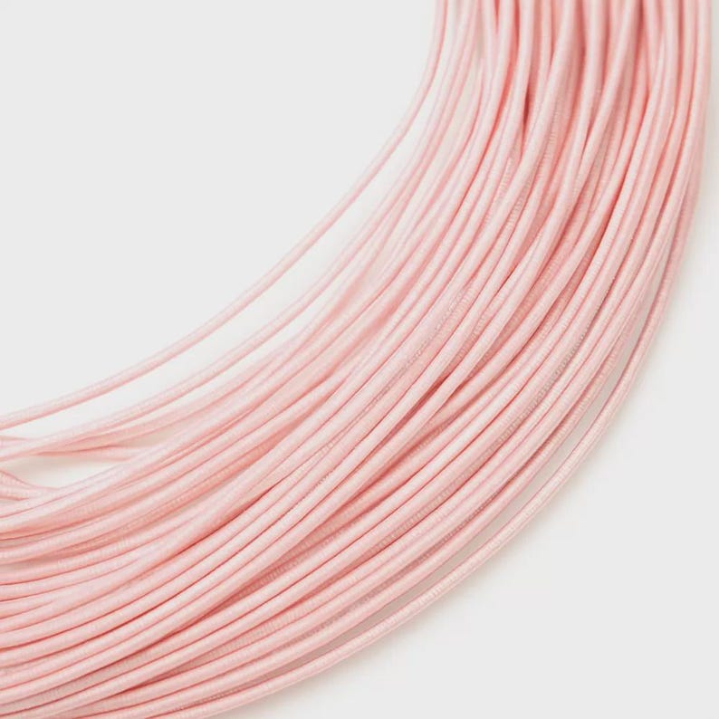 May include: A collection of thin, light pink flexible cords arranged in a curved, layered formation. The individual strands feature a smooth, uniform texture and a consistent pastel pink hue, creating a dense, flowing pattern of parallel lines.